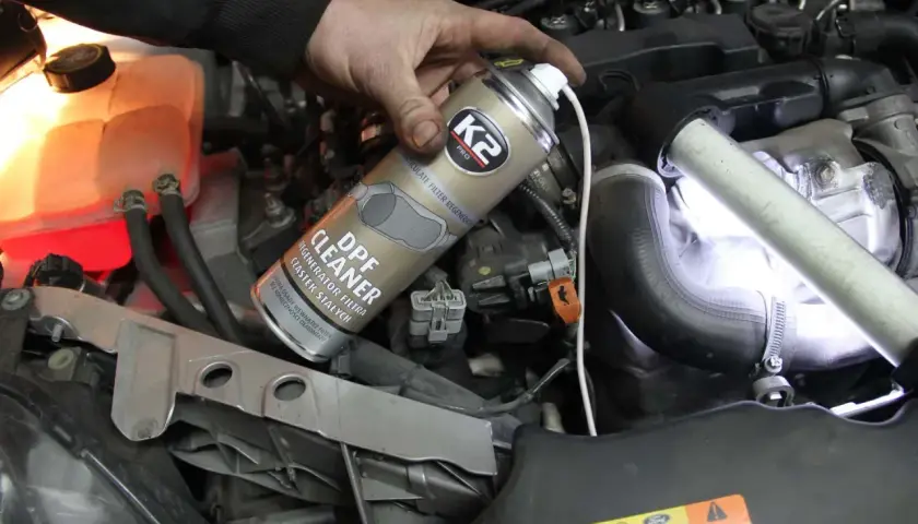 Ręka trzymająca puszkę preparatu do czyszczenia DPF na tle silnika samochodowego.