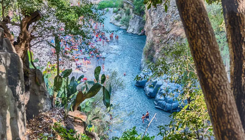 Gente si gode il sole e l'acqua cristallina nella Valle dell'Alcantara, tra rocce vulcaniche e vegetazione rigogliosa.