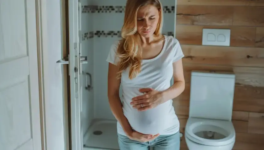 Kobieta w ciąży z wyrazem bólu na twarzy trzyma się za brzuch. W tle łazienka z toaletą. Ból podbrzusza w ciąży.