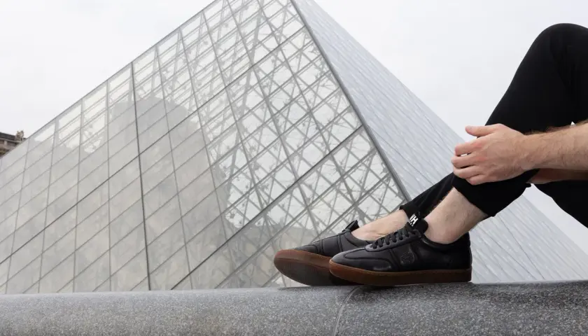 Un homme porte une tenue avec basket noir, assis devant la pyramide du Louvre.