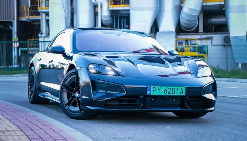 Elegancki Porsche Taycan w ciemnym kolorze, zaparkowany przed industrialnym budynkiem z rurami.