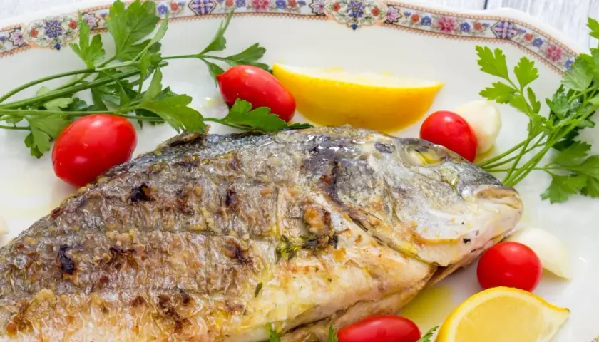 Orata alla griglia dorata e succulenta, guarnita con pomodorini, spicchi d'aglio, limone e prezzemolo fresco.