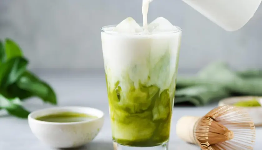 Milch wird in ein Glas mit Eis und Matcha gegossen. Ein Löffel mit Matcha-Pulver und eine Schale mit Matcha-Tee sind daneben zu sehen. Ein traditioneller Matcha-Besen liegt auf dem Tisch.