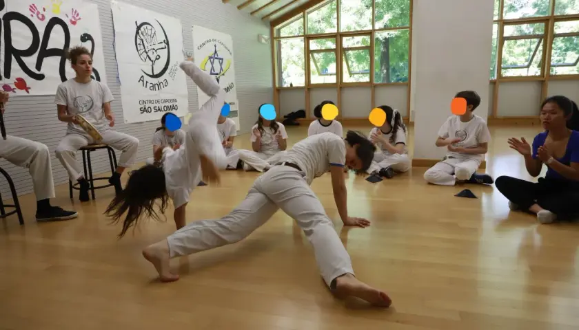 Bambini praticano capoeira in una palestra luminosa. Uno fa una capriola, altri osservano e applaudono.
