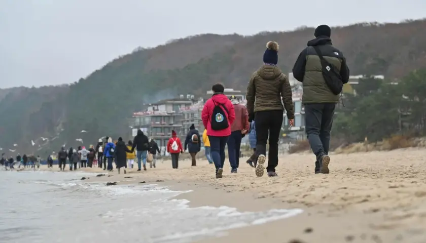 Spacer po plaży, gdy jest długi weekend. Ludzie w ciepłych ubraniach idą brzegiem morza, w tle widać zabudowania i wzgórza.