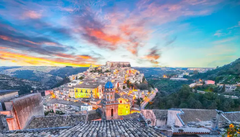 A picturesque Italian town perched on a hill, bathed in the warm glow of sunset. This view captures the essence of **two weeks in Sicily**, with its charming architecture and vibrant sky.