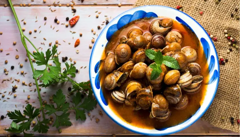 Un plato de caracoles en salsa, adornado con menta fresca, rodeado de especias y perejil sobre una mesa de madera.
