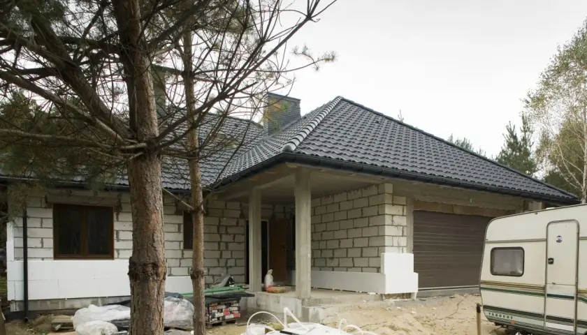 Budowa domu w toku. Widać białe płyty styroduru przy ścianach, które będą izolacją. Obok stoi przyczepa kempingowa.
