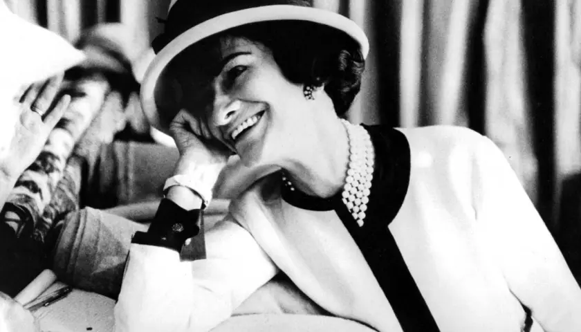 Coco Chanel, sonriendo con un sombrero y perlas, icónica figura de la moda.