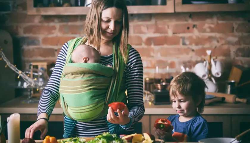 Mama z dzieckiem w chuście i synem przygotowują posiłek. Od kiedy można nosić dziecko w chuście? Od pierwszych dni życia!