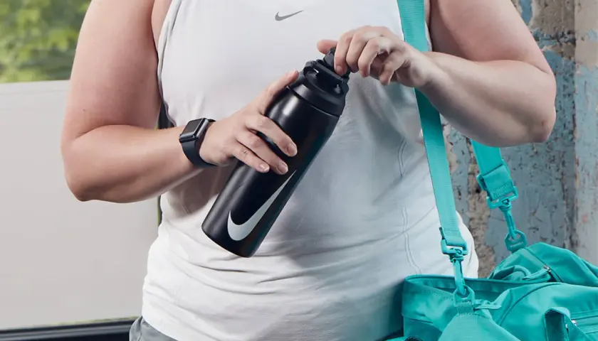 Osoba w białej koszulce sportowej trzyma czarną butelkę Nike. Obok wisi turkusowa torba.