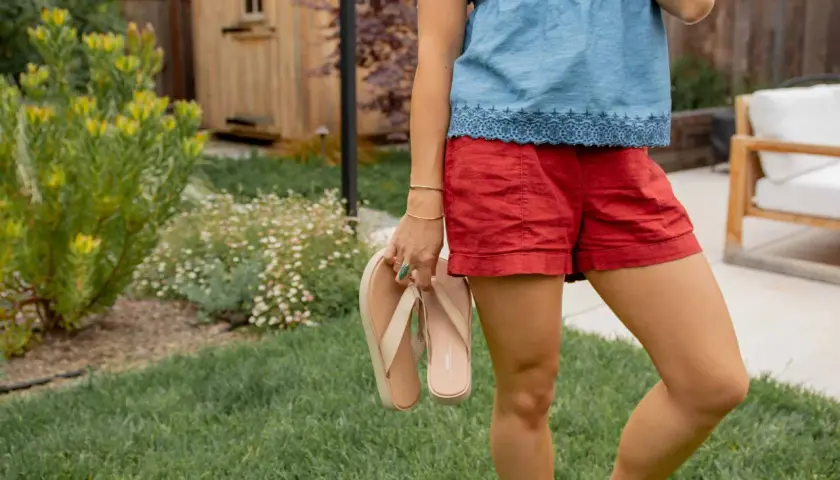 Mujer con blusa vaquera y shorts rojos, sosteniendo chanclas. El jardín tiene césped y flores.