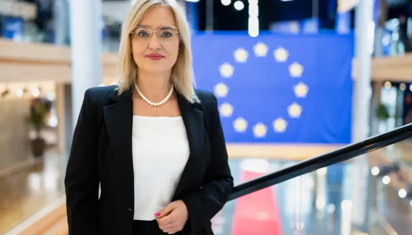 Magda Adamowicz, w eleganckim stroju, stoi przed flagą Unii Europejskiej. Jej wiek nie jest widoczny, ale emanuje pewnością siebie.