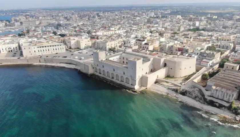 Vue aérienne du château de Trani, une forteresse médiévale sur la côte, avec la ville en arrière-plan et la mer d'un bleu-vert éclatant.