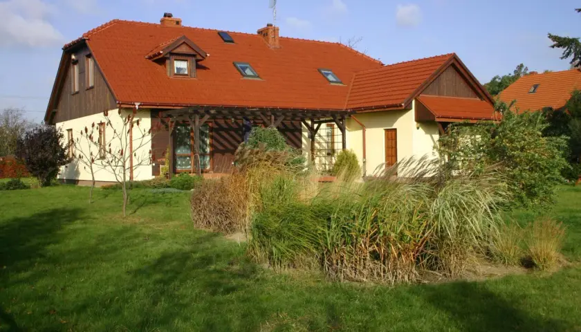 Dom z czerwonym dachem naczółkowym, z werandą i ogrodem pełnym traw ozdobnych.