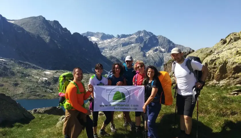 Grupo de excursionistas sonríe en la montaña, listos para la ruta. Llevan mochilas y un cartel que dice "Aquí empieza tu viaje".