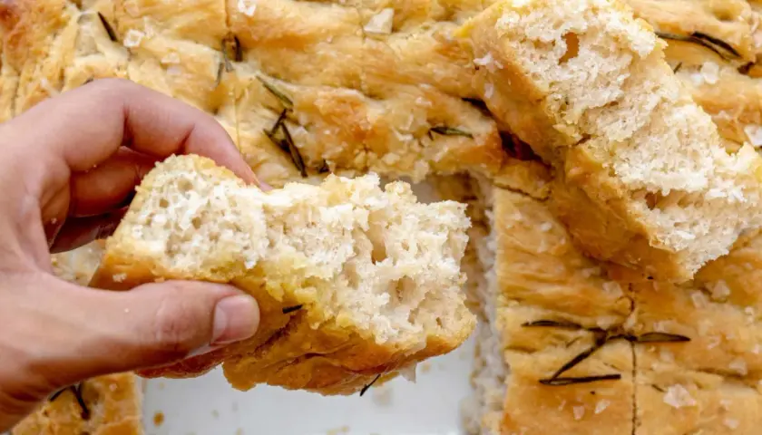 Mano rompe un trozo de focaccia original, dorada y con romero.