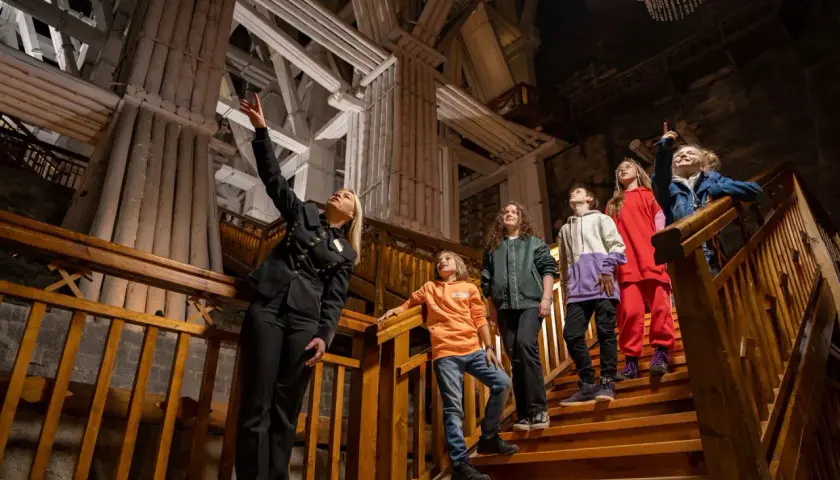 Przewodniczka oprowadza grupę dzieci po Kopalni Soli "Wieliczka". Wszyscy są ubrani na cebulkę, co sugeruje stałą, niską temperaturę w kopalni.