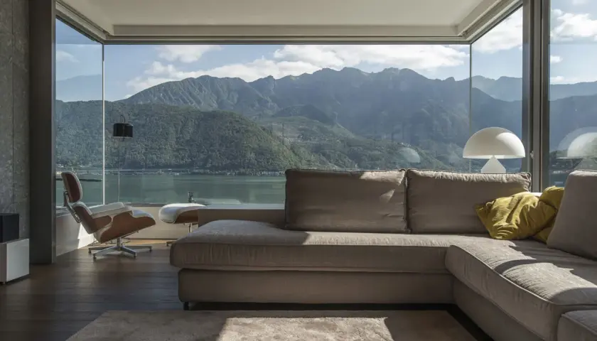 Soggiorno moderno con vista panoramica su lago e montagne, reso possibile da ampi infissi di ultima generazione. Divano, poltrona e tappeto creano un'atmosfera accogliente.
