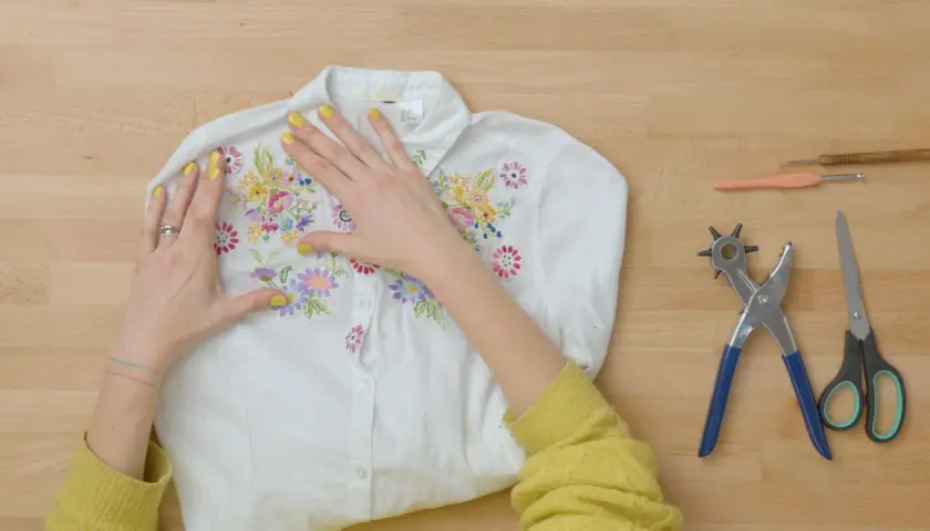 Manos embelleciendo una camisa blanca con bordados florales, lista para un proyecto de upcycling ropa. Herramientas de costura a un lado.