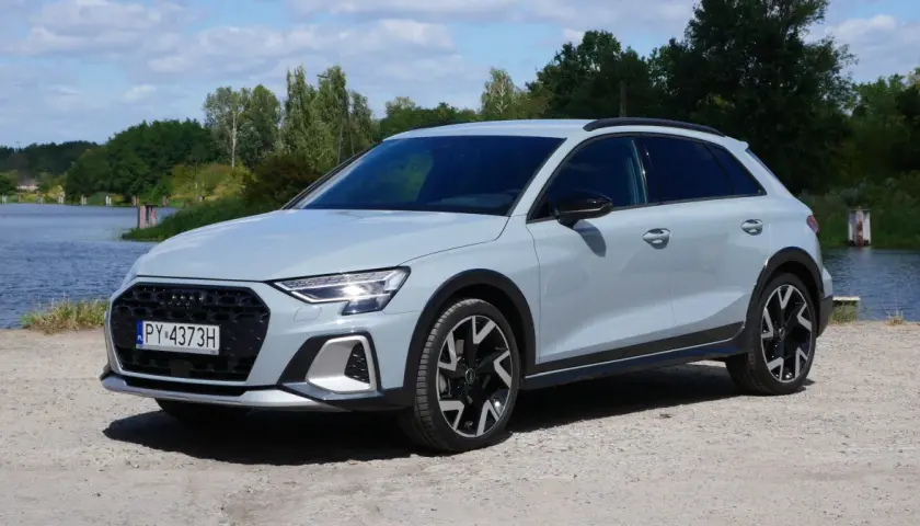 Jasnoszary Audi A3 Allstreet z czarnymi dodatkami stoi na żwirowej drodze nad jeziorem.
