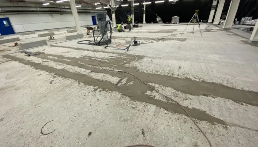 Prace przy naprawie posadzki betonowej. Widać sprzęt budowlany, pracowników i przygotowane do wylania fugi.