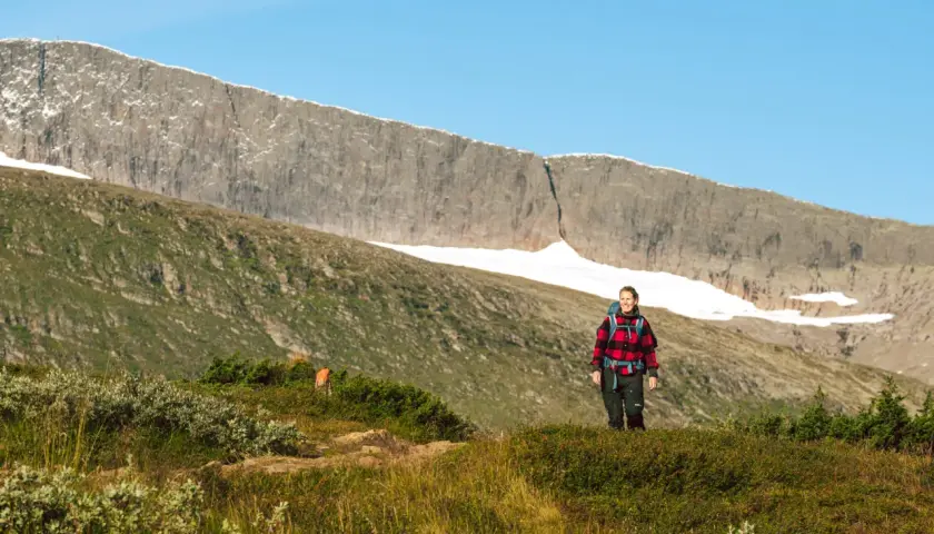 En kvinna på helags topptur njuter av utsikten över fjällmassivet. Solen lyser på den gröna sluttningen.