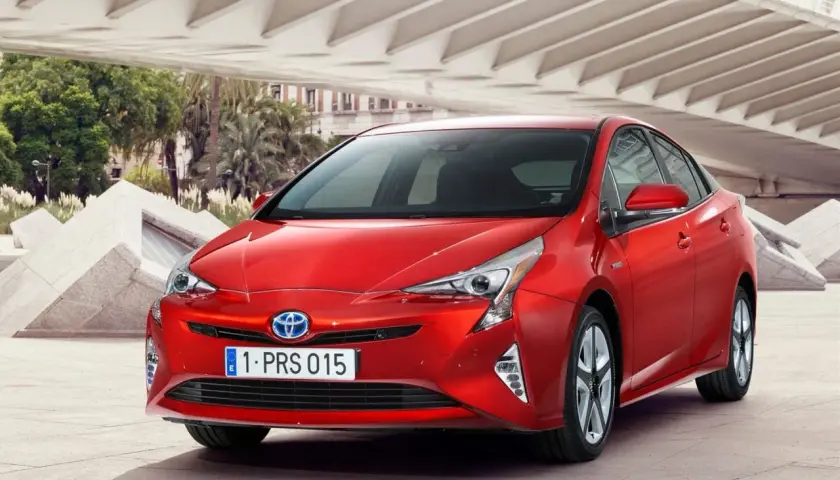 Czerwony Prius 4 z nowoczesnym designem, zaparkowany pod betonową konstrukcją z palmami w tle.