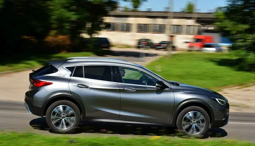 Srebrny Infiniti QX30 pędzi po drodze, mijając zielone pobocze i budynki.