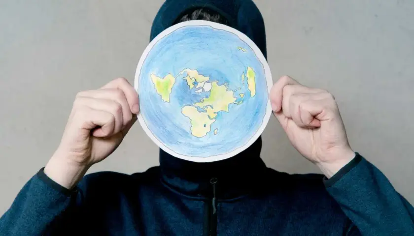 Osoba w kapturze trzyma okrągły rysunek płaskiej Ziemi. To nie jest globe po polsku, ale ciekawa wizja świata.