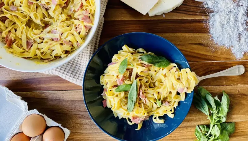 Un plat de pâtes à la carbonara crémeuses, garni de basilic frais. Des ingrédients comme des œufs, du fromage et de la farine sont visibles à proximité.