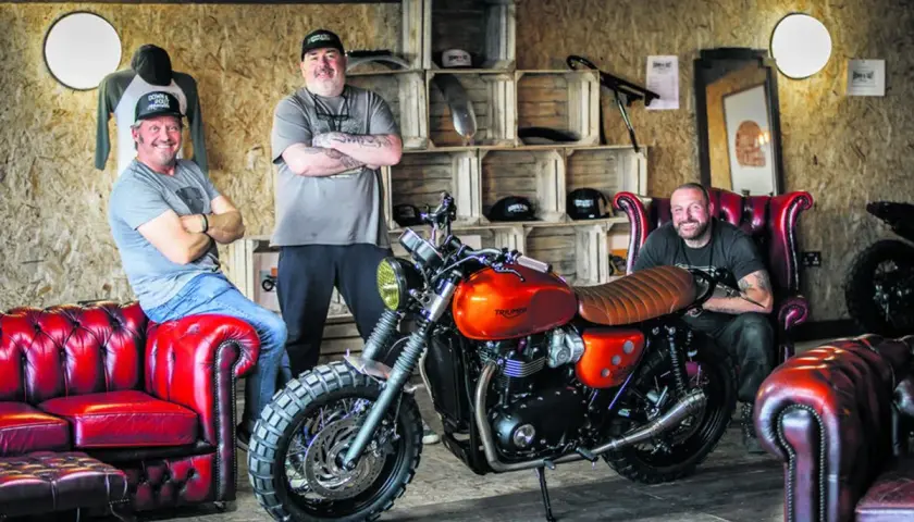 Three men pose with a custom orange Triumph motorcycle, showcasing the work of best custom bike builders.