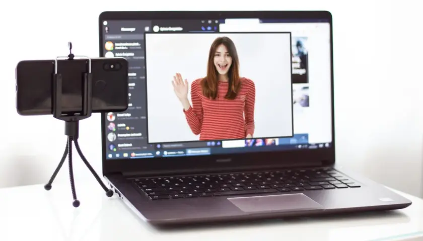 Kamera z telefonu na PC: młoda kobieta w bluzce w paski wita się na ekranie laptopa, obok stoi telefon na statywie.