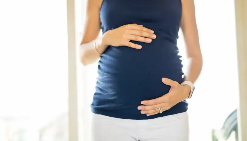 Ciężarna kobieta w granatowej bluzce i białych spodniach obejmuje dłońmi brzuch. Wody płodowe otulają rozwijające się życie.