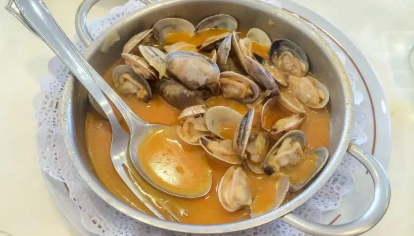 Almejas en una cazuela metálica, bañadas en una sabrosa salsa marinera. Una cuchara plateada reposa sobre el plato, lista para disfrutar.