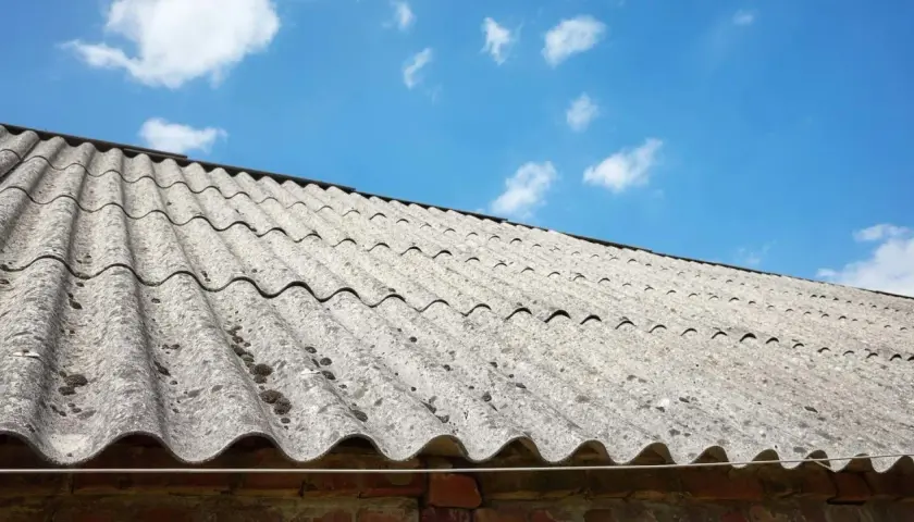 Stary dach pokryty eternitem, materiałem zawierającym azbest, pod błękitnym niebem z chmurami.