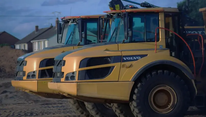Dwa żółte wywrotki Volvo A30D stoją obok siebie. W tle domy i niebo.