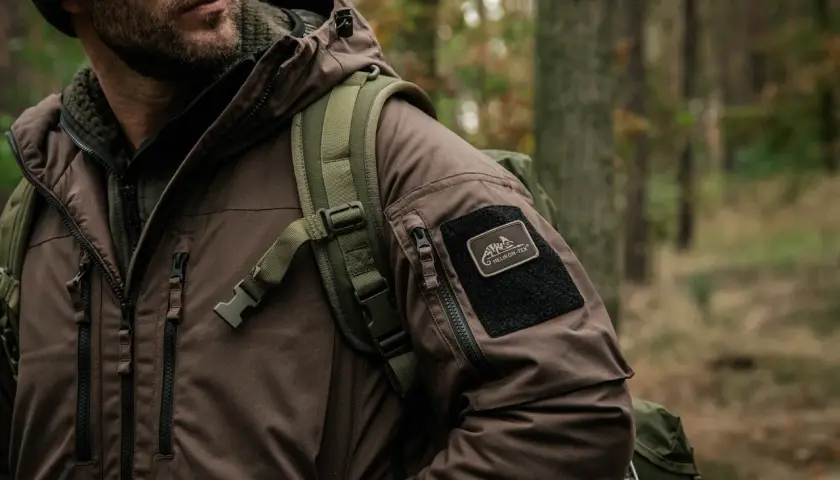 Mężczyzna w brązowej kurtce z naszywką Helikon-Tex, symbol polskie marki sportowe, w lesie.