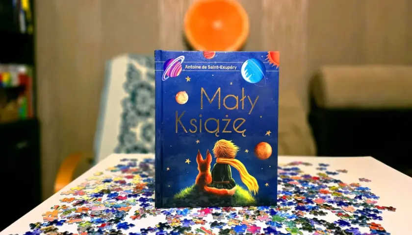 Odkryj, dlaczego warto przeczytać Małego Księcia. Książka z liskiem i chłopcem na tle kosmosu, otoczona rozsypanymi puzzlami.