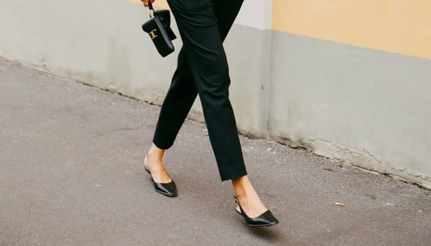 Une femme élégante marche dans la rue, portant un pantalon noir et des chaussures ballerines noires. Elle tient un petit sac noir.