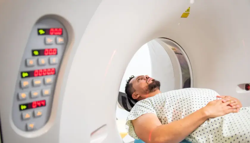 Mężczyzna leży w aparacie do tomografii komputerowej, przygotowując się do badania. Obok widoczny panel sterowania z cyfrowymi wskaźnikami.