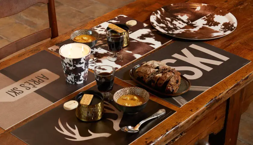 Ambiance chaleureuse pour une deco table raclette : bougie, bols de soupe, pain et verres de vin sur des sets "après ski" et motif vache.