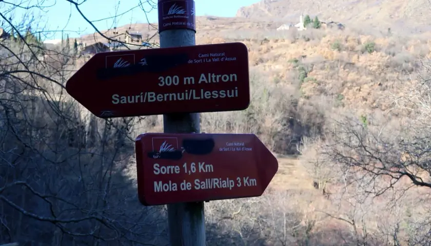 Señales de senderismo en la Vall d'Assua: 300m a Altron y 1,6km a Sorre. El paisaje montañoso y boscoso sugiere una ruta de naturaleza.
