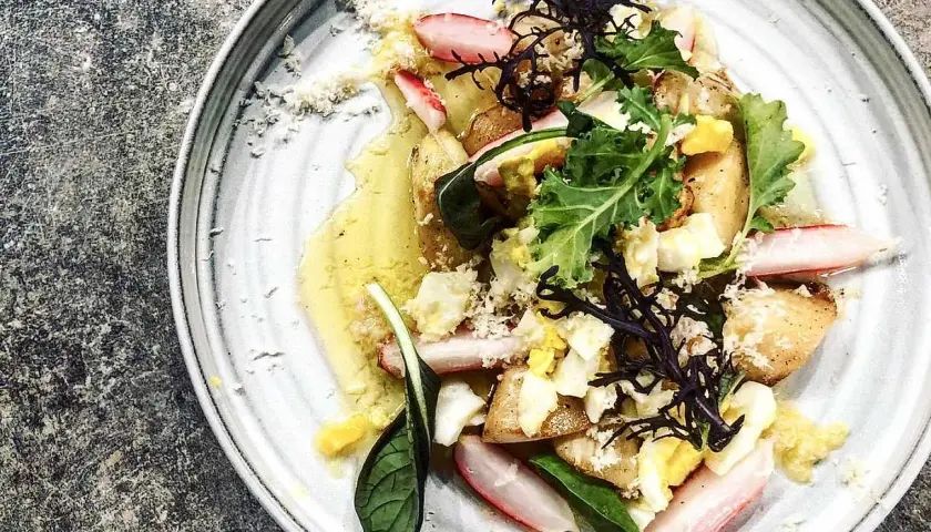 En vackert presenterad rätt med potatis, ägg, gröna blad och skivor av vit sparris, dränkt i en gyllene sås.
