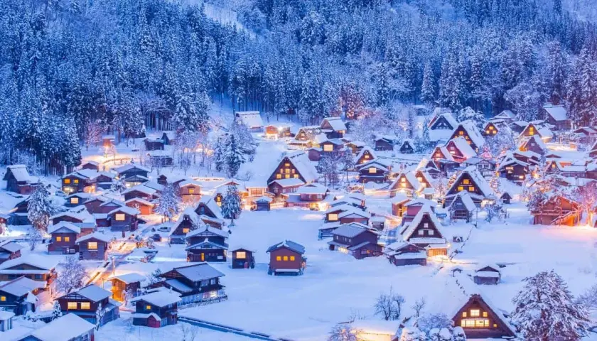 Ein verschneites Dorf in Japan im Winter. Traditionelle Häuser mit warmem Licht sind von schneebedeckten Bäumen umgeben.