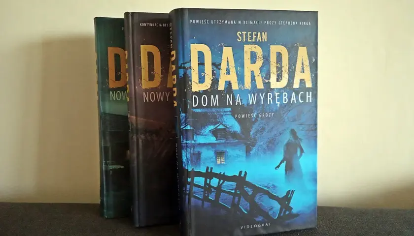 Trzy książki Stefana Dardy, w tym "Dom na wyrębach", stoją obok siebie. Okładka "Domu na wyrębach" przedstawia mroczny, niebieski krajobraz z domem i postacią.