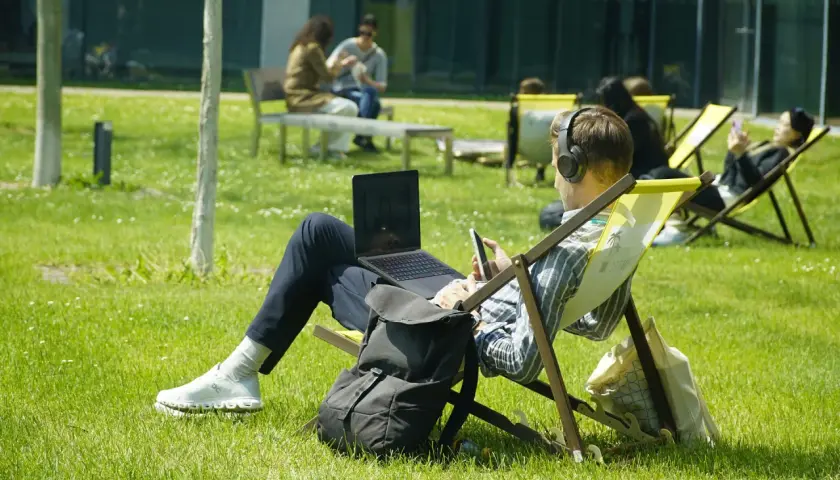 Mann mit Kopfhörern und Laptop im Freien, genießt sein Fernstudium im Ausland auf einer Liege im Sonnenschein.
