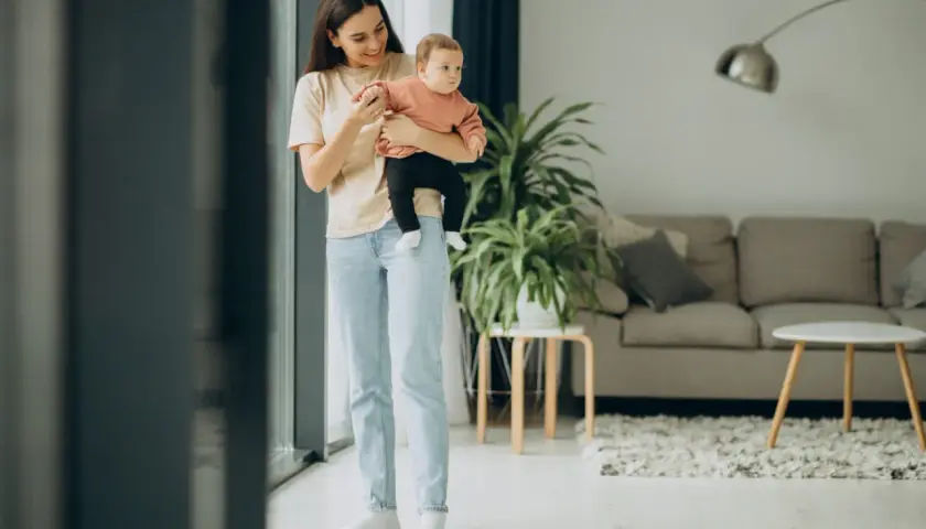 Uśmiechnięta kobieta z dzieckiem na rękach stoi przy oknie w jasnym salonie.