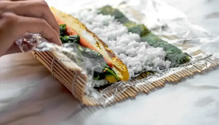Ręce zwijają domowe sushi na macie bambusowej. Ryż, nori, omlet i warzywa tworzą pyszne wnętrze rolki.