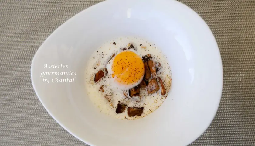 Un œuf poché, entouré de morceaux de champignons et d'une mousse légère, dans une assiette blanche. Des oeufs parfaits pour un repas gourmand.